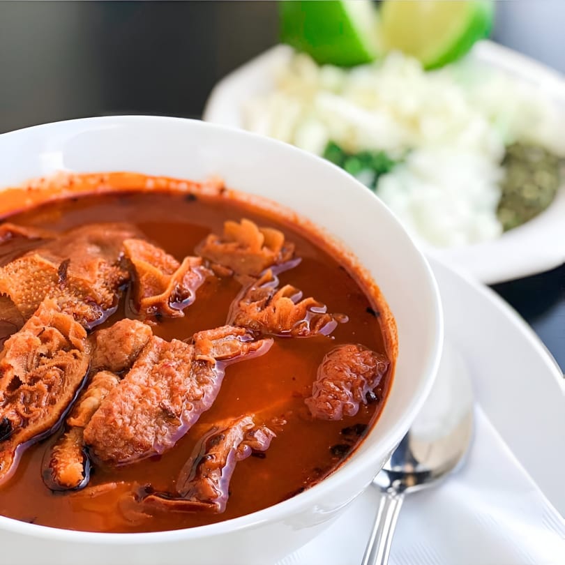 Menudo soup in a white bowl with a side of chopped onions, cilantro, and lime.