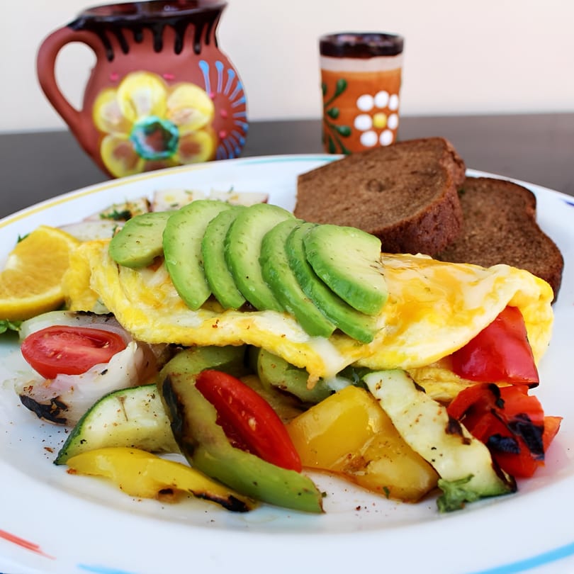 A colorful omelet topped with avocado slices, grilled vegetables, and a side of dark bread.